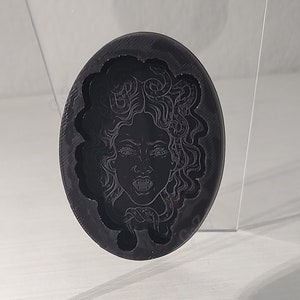 May include: A black circular resin mold with a detailed design of a woman's face with snakes in her hair. The mold is on a clear acrylic display stand.