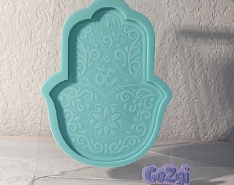 May include: A teal blue silicone mold of a hamsa hand with an intricate floral design. The mold is on a clear acrylic stand with the word "Go2gi" printed on it.