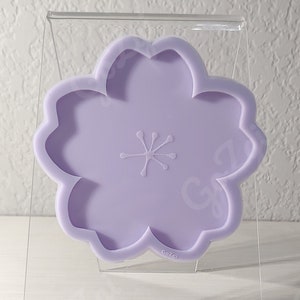 May include: A light purple silicone mold shaped like a flower with five petals. The mold is on a clear acrylic stand.