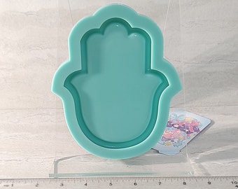 May include: A teal blue silicone mold shaped like a hamsa hand. The mold is in a clear plastic package. A gray silicone mold of the same shape is shown next to the package.