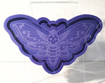 May include: Purple silicone mold of a moth with a skull design. The mold is on a clear acrylic stand.