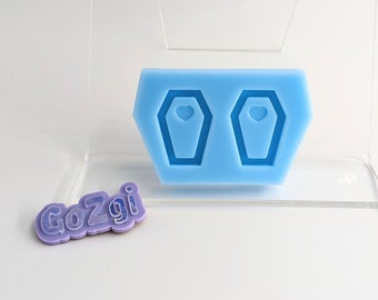 May include: A light blue silicone mould featuring two coffin shapes with heart details. A small, lavender-coloured emblem with the text "GoZgi" is also present. The mould is displayed on a clear acrylic stand against a white background.