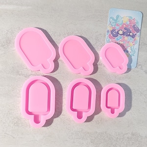 Może przedstawiać: Sześć różowych silikonowych form w kształcie lodów na patyku w różnych rozmiarach. Formy znajdują się na jasnoszarej, teksturowanej powierzchni.