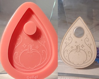 May include: A light orange silicone mould and a wooden planchette, both teardrop-shaped. The mould has an embossed pumpkin design with stars and swirls. The wooden planchette features a pumpkin design and a circular opening.