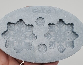 Op de afbeelding: Een grijze siliconen mal met twee sneeuwvlokontwerpen. De mal is ovaalvormig en heeft het woord "Gozgi" erop gedrukt.
