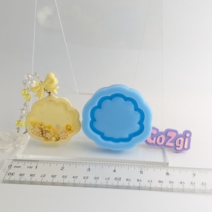 May include: A blue silicone mold shaped like a shell with a raised edge. A clear resin pendant with a yellow bow and gold glitter is next to the mold. A ruler is in the background with the word "CoZgi" printed on it.