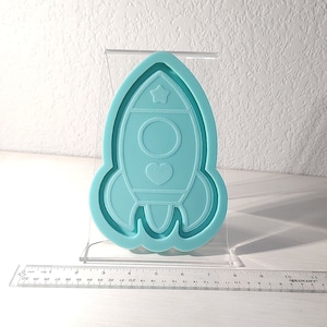 Puede incluir: Un molde de silicona azul claro con forma de cohete con una estrella en la parte superior y un corazón en el medio. El molde está en un soporte de acrílico transparente.