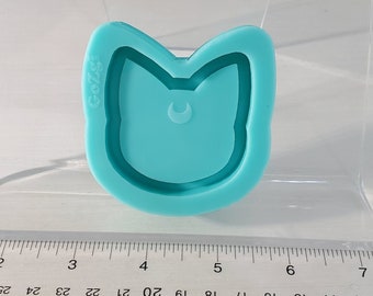 May include: A turquoise silicone mould shaped like a cat's face with a crescent moon inside. The mould is on a white surface with a ruler.