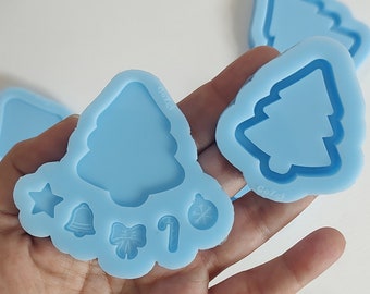 May include: Light blue silicone molds in various Christmas shapes, including a Christmas tree, star, bell, bow, candy cane, and ornament. The molds are held in a hand against a white background. These molds are for crafting.