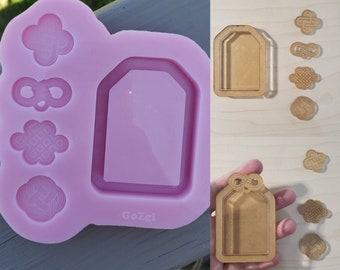 Puede incluir: Un molde de silicona rosa con seis formas diferentes, incluyendo una etiqueta, un nudo y un corazón. El molde está etiquetado como "GoZgi". También hay seis recortes de madera de las mismas formas sobre una superficie de madera clara.