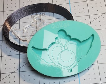 May include: A teal silicone mould featuring a heart and wing design, with a black plastic ring and a clear plastic shape. The mould has the text "GoZgi" printed on it. The items are on a grid-patterned surface.