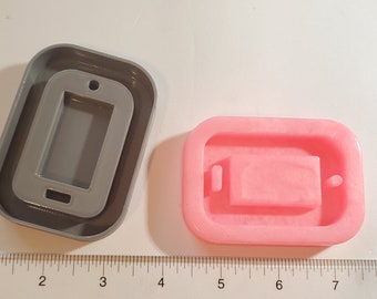 Puede incluir: Dos moldes de silicona para manualidades, uno gris y otro rosa. El molde gris tiene forma rectangular con un recorte en forma de teléfono. El molde rosa tiene forma rectangular con un recorte en forma de batería. Una regla está colocada junto a los moldes.