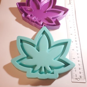 Puede incluir: Dos moldes de silicona en forma de hoja de marihuana. El molde más grande es morado y el más pequeño es verde azulado.