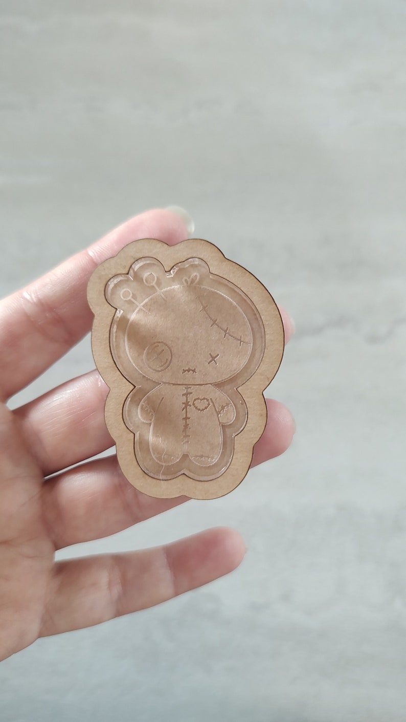 May include: A wooden cutout of a stitched doll with a heart on its chest. The doll has a stitched-up mouth and eyes, and a stitched-up body. The cutout is brown and has a white outline.