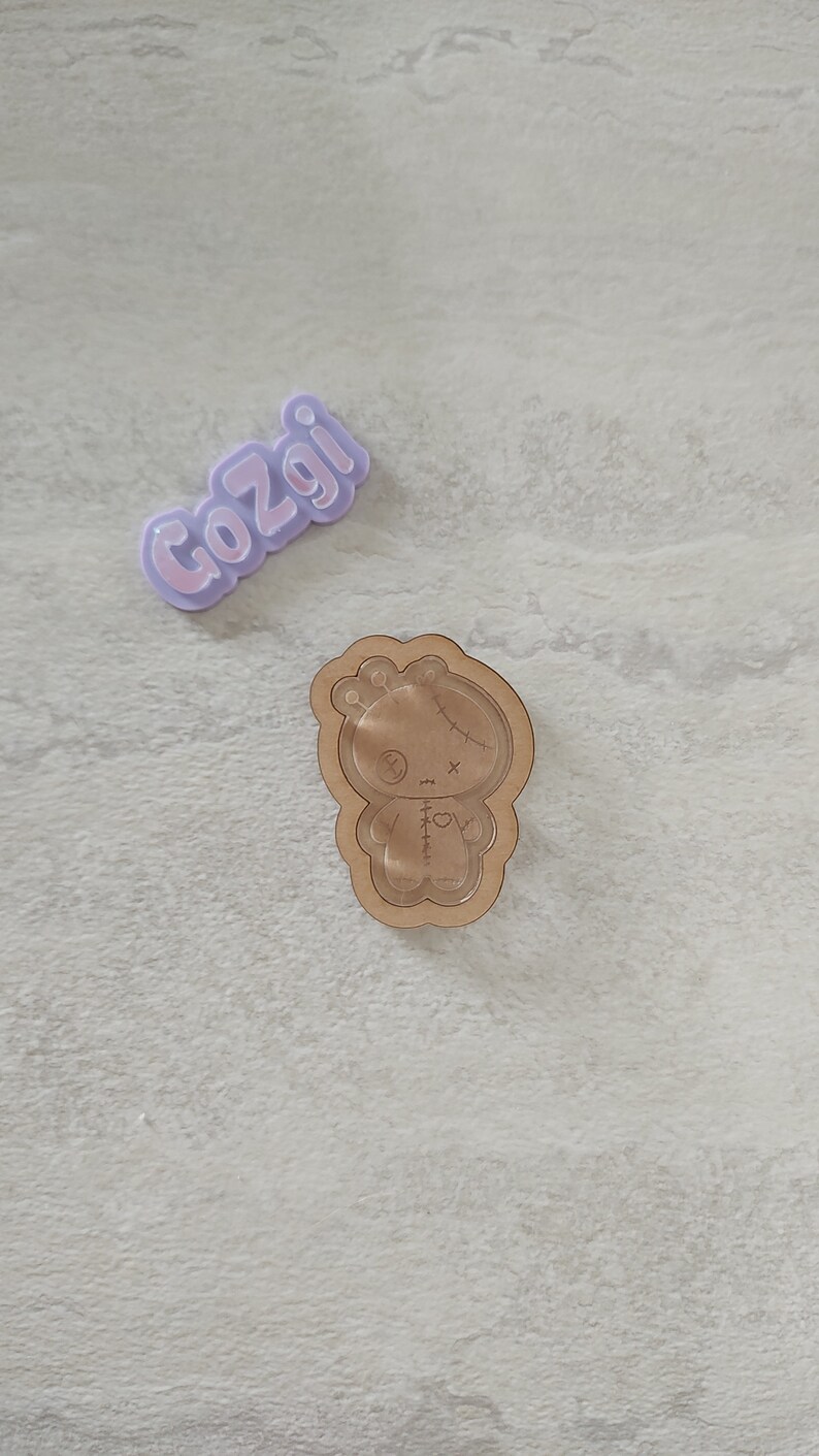 May include: A wooden cutout of a stitched doll with a heart on its chest. The doll has a stitched X for eyes and a stitched mouth. The cutout is on a light gray background.