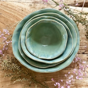 May include: A set of three nested ceramic bowls with a glossy, light sage green glaze. Each bowl features an organic, wavy, and uneven rim design. The bowls are stacked inside one another, showcasing a smooth, reflective surface and a consistent color palette throughout the set, creating a cohesive appearance for serving or display purposes.