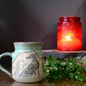 May include: A handmade ceramic mug with a wolf design, featuring a light green rim and handle. Beside it, a red glass candle holder with a lit candle sits on a wooden shelf. Greenery adds a natural touch.