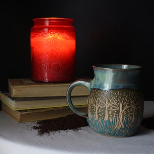 May include: A ceramic mug with a tree design, a red candle in a jar, and a stack of books. The mug has a blue and green glaze. The candle is lit, casting a warm glow. The scene is set on a white tablecloth.