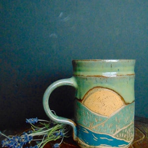 May include: A ceramic mug with a green, blue, and yellow landscape design. The mug has a handle and is sitting on a wooden surface.