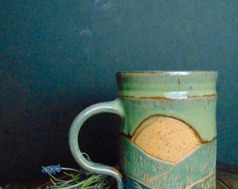 May include: A ceramic mug with a green, blue, and yellow landscape design. The mug has a handle and is sitting on a wooden surface.