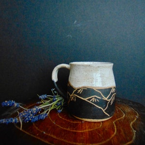 May include: A white ceramic mug with a black and brown mountain design. The mug has a speckled glaze and a rounded shape. It is sitting on a wooden surface with a sprig of lavender.