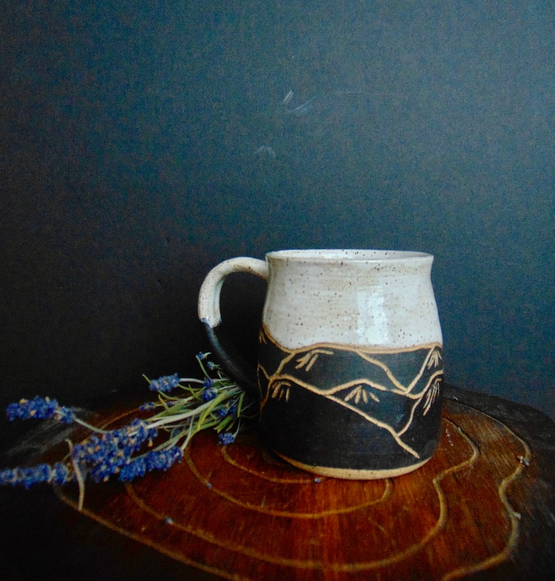 K&ouml;nnte beinhalten: Eine wei&szlig;e Keramiktasse mit einem schwarzen und braunen Bergdesign. Die Tasse hat eine gesprenkelte Glasur und eine abgerundete Form. Sie steht auf einer Holzoberfl&auml;che mit einem Lavendelzweig.