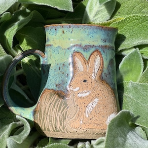 May include: A handmade ceramic mug with a rabbit design. The mug features a brown rabbit on a background of green and blue glaze. The handle is dark brown. The mug is surrounded by green leaves.