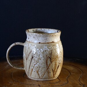 May include: A handmade ceramic mug with a beige and white speckled finish. The mug features a curved handle and is decorated with an embossed floral design. The mug is approximately 4 inches tall and is suitable for hot or cold beverages.