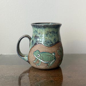 May include: Handmade ceramic mug with a green and blue speckled glaze. The mug features a hand-painted green frog design with white accents. The handle is a matching blue-green color. The mug is a unique piece of pottery.