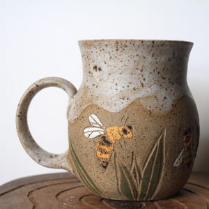 May include: A handmade ceramic mug with a beige and brown speckled finish. The mug features a hand-painted design of a bee with yellow and black details, along with green grass. The mug has a curved handle and is suitable for hot beverages.