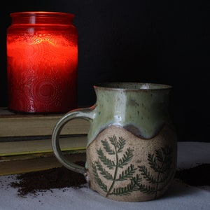 May include: A handmade ceramic mug with a green and brown glaze, featuring a fern leaf design. A red glass candle holder sits behind the mug, next to a stack of books. The scene is set on a white surface with a dark background.