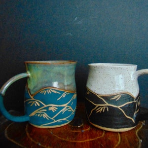 K&ouml;nnte beinhalten: Zwei Keramikbecher mit einem Bergketten-Design. Ein Becher ist blau und gr&uuml;n mit goldenen Akzenten, der andere ist schwarz und wei&szlig; mit goldenen Akzenten.