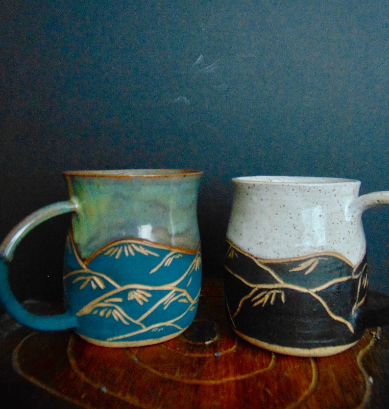 K&ouml;nnte beinhalten: Zwei Keramikbecher mit einem Bergketten-Design. Ein Becher ist blau und gr&uuml;n mit goldenen Akzenten, der andere ist schwarz und wei&szlig; mit goldenen Akzenten.
