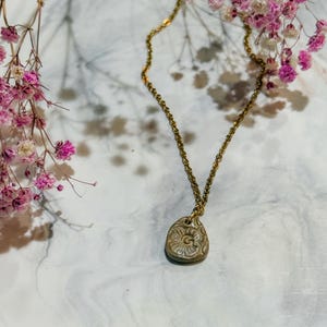 Könnte beinhalten: Goldfarbene Halskette mit Anhänger. Der Anhänger hat eine Tropfenform mit floralem Design. Die Halskette liegt auf einer Marmoroberfläche mit rosa Blüten.