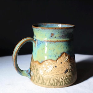 May include: A handmade ceramic mug with a deer design. The mug features a light blue and green glaze on the upper portion, transitioning to a tan base with an etched deer and grass design. The handle is curved and matches the glaze.