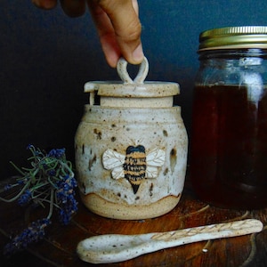 May include: A ceramic honey pot with a bee design, a matching spoon, and a jar of honey. The honey pot is beige with a bee applique. The spoon and pot have a speckled finish. A hand is lifting the lid of the honey pot.
