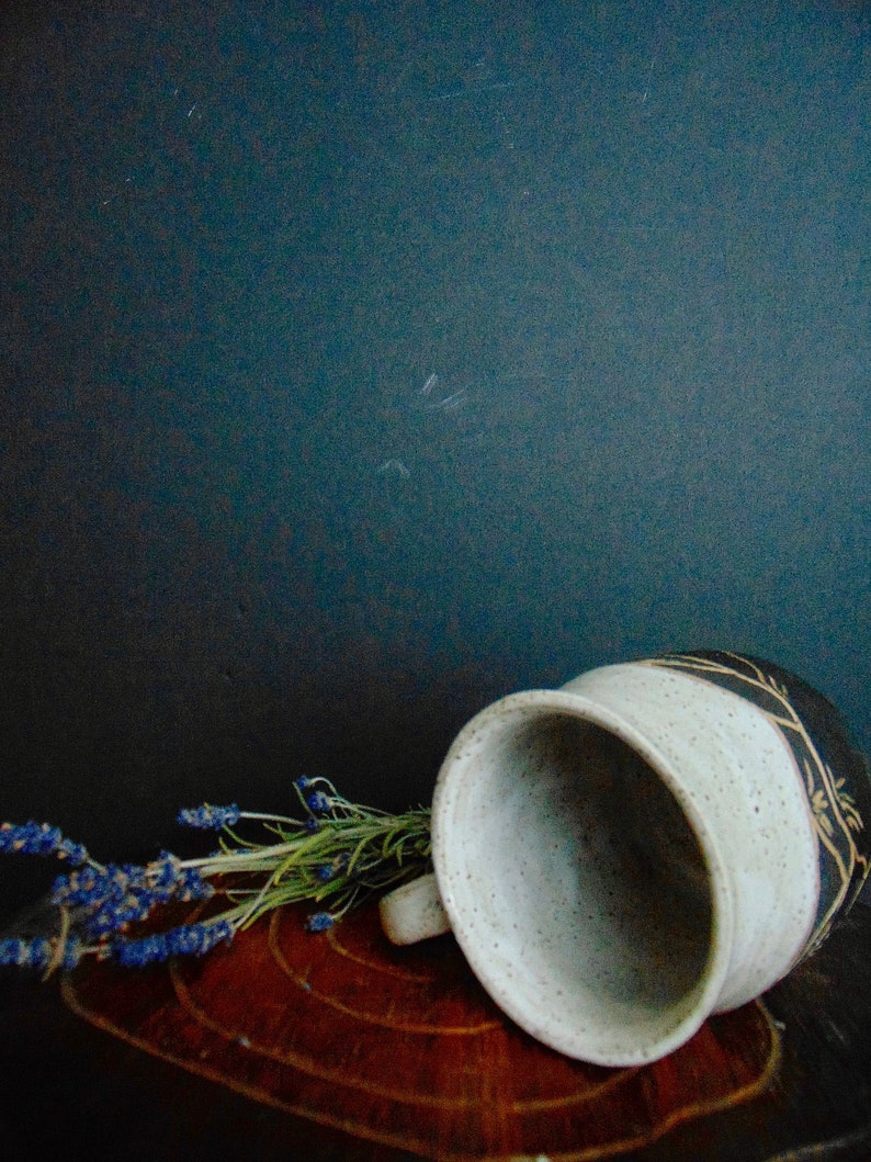 K&ouml;nnte beinhalten: Eine wei&szlig;e Keramiktasse mit einem schwarzen und goldenen Design liegt auf einer Holzoberfl&auml;che. Ein Lavendelzweig liegt neben der Tasse.