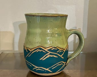 May include: A green ceramic mug with a blue and brown mountain design. The mug has a rounded body and a handle.