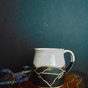 K&ouml;nnte beinhalten: Eine wei&szlig;e Keramiktasse mit einem schwarzen und braunen Bergmotiv. Die Tasse hat eine gesprenkelte Glasur und einen abgerundeten Griff.