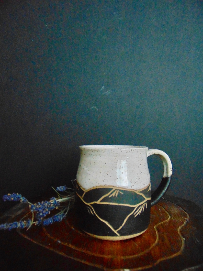 K&ouml;nnte beinhalten: Eine wei&szlig;e Keramiktasse mit einem schwarzen und braunen Bergmotiv. Die Tasse hat eine gesprenkelte Glasur und einen abgerundeten Griff.