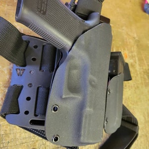 May include: A black leather holster with a black pistol inside. The holster is attached to a black tactical belt with a strap that goes over the shoulder.