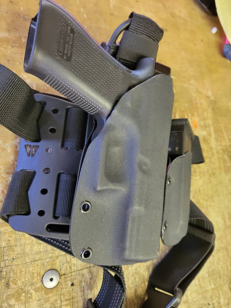 May include: A black leather holster with a black pistol inside. The holster is attached to a black tactical belt with a strap that goes over the shoulder.