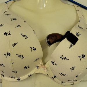May include: A white bra with a floral pattern and a small black gun holster attached to the strap. A small handgun is in the holster.