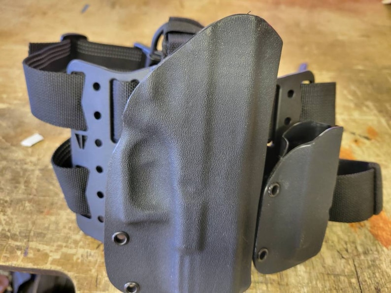 May include: Black tactical leg holster with adjustable straps and multiple attachment points. The holster is made of durable plastic and has a smooth, matte finish.