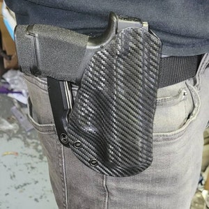 May include: A black handgun in a carbon fiber pattern holster attached to a black belt. The holster is worn on the right side, secured to the waist. The person is wearing gray jeans and a black shirt.