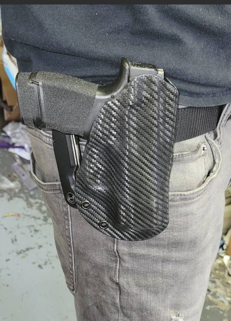 May include: A black handgun in a carbon fiber pattern holster attached to a black belt. The holster is worn on the right side, secured to the waist. The person is wearing gray jeans and a black shirt.