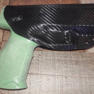 Puede incluir: Una pistola negra y verde con una funda de fibra de carbono. La funda tiene tres pequeños agujeros en el lateral.