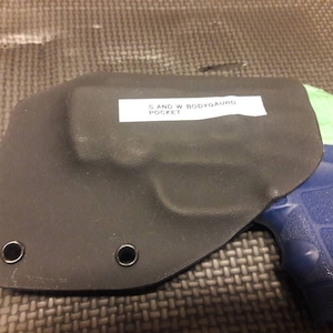 Puede incluir: Una funda de cuero negra con dos ojales metálicos y una etiqueta que dice "S and W Bodyguard Pocket". Una pistola azul y verde es parcialmente visible en la funda.