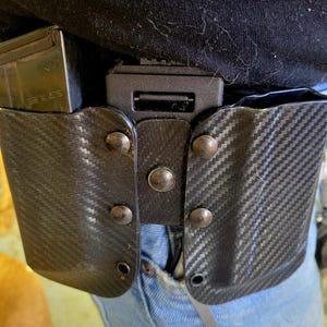 May include: A black carbon fiber holster system with a magazine pouch attached to a belt. The holster is secured with rivets and a black buckle. The magazine is marked with "5.7 x 28".