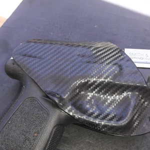 May include: A black handgun with a carbon fiber holster. The holster has a textured surface and is designed to fit snugly around the gun. The text "SMITH & WESSON" is visible on the holster.